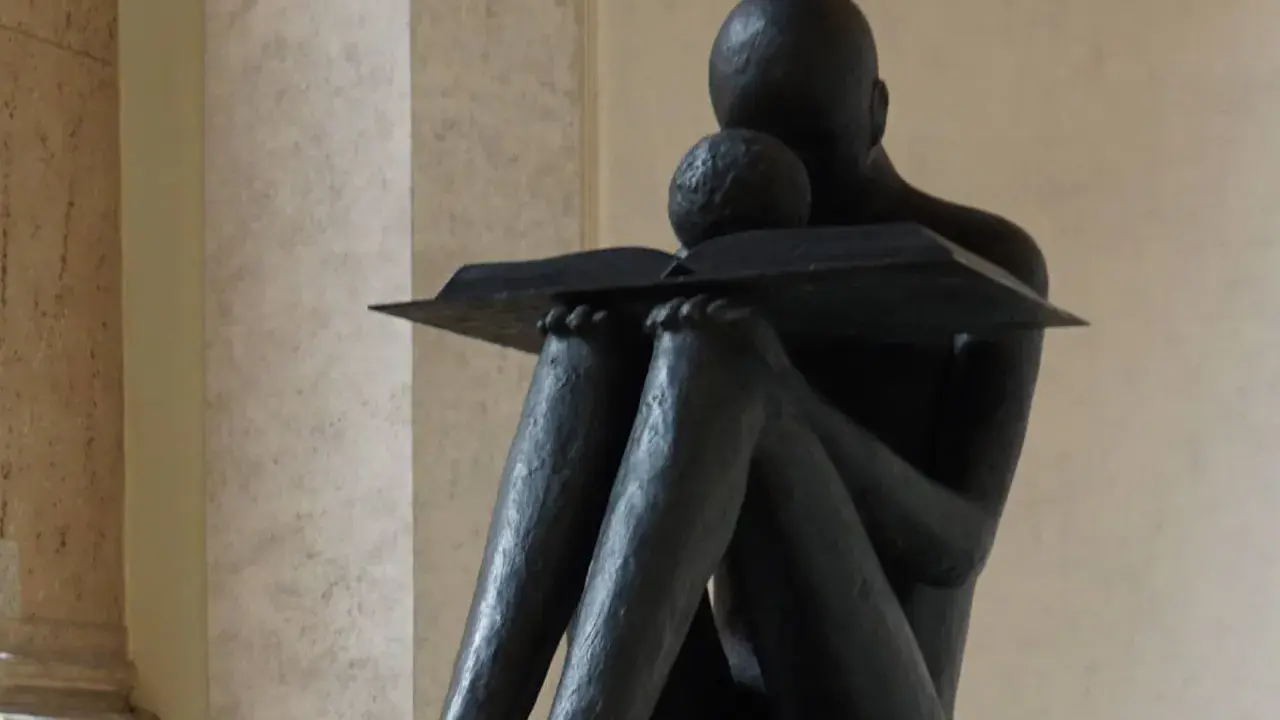 Statua di una figura che legge un libro, con un bambino appoggiato, immersi nella conoscenza a tutto spiano.