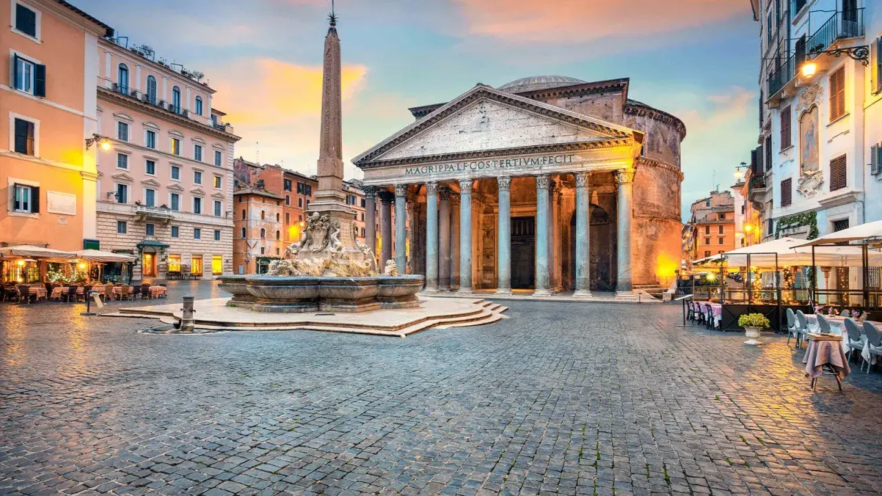 Piazza illuminata con il Pantheon, una chiesa maestosa, e una fontana con obelisco. Tavolini all'aperto invitano a godersi la sera romana.