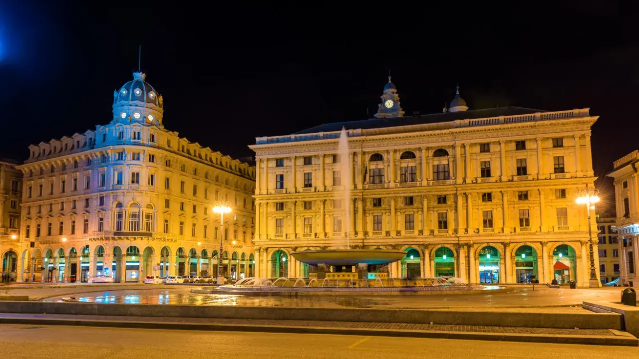 Piazza De Ferrari illuminata di notte, con la fontana centrale e gli edifici storici che animano la vita notturna di Genova.