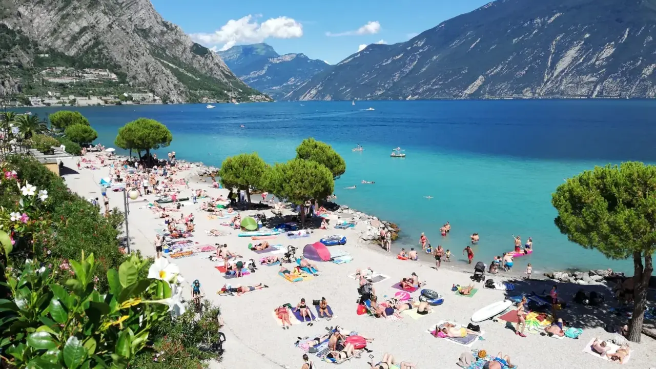 Spiaggia di Limone sul Garda affollata di bagnanti sotto le montagne. Acqua cristallina e sole splendente.