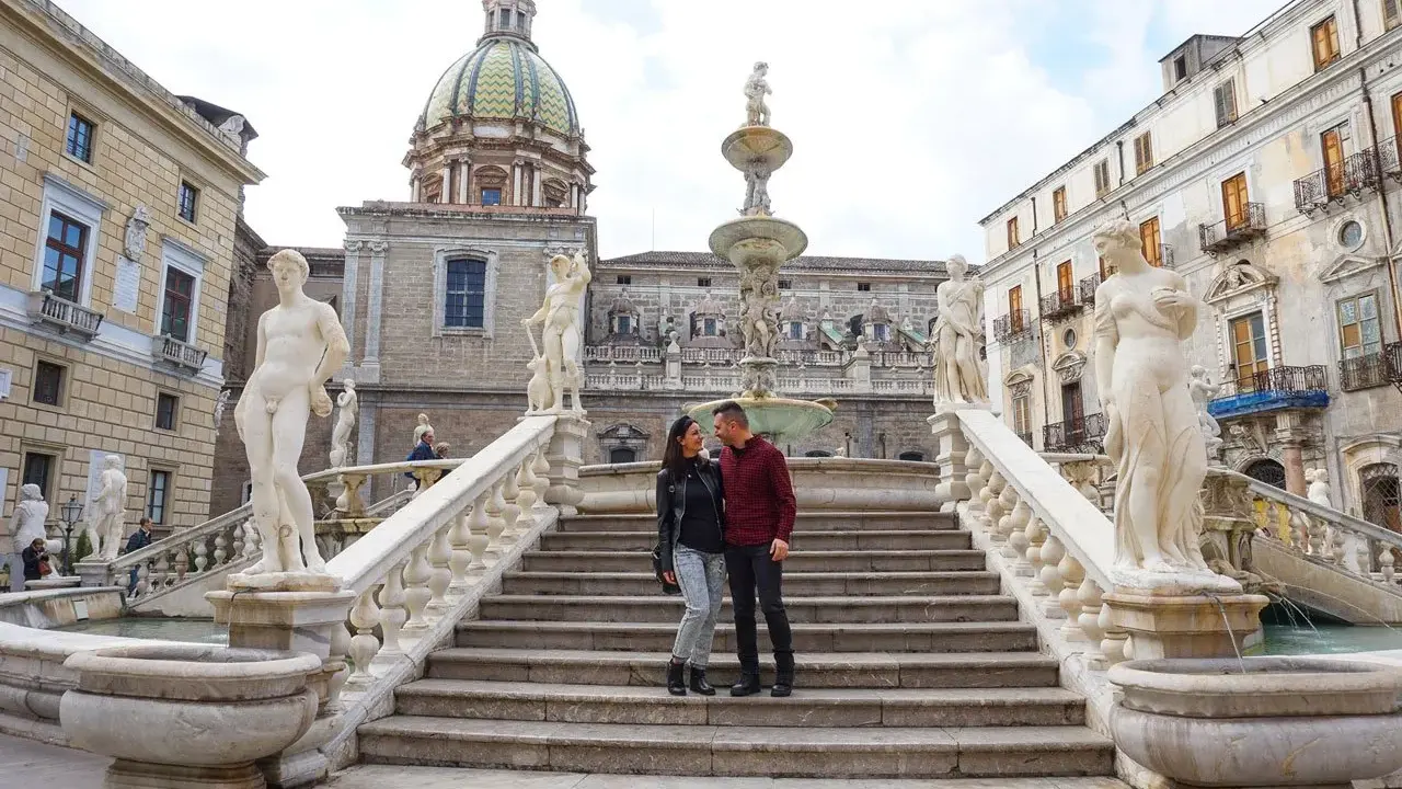 Coppia felice davanti alla Fontana Pretoria, un luogo imperdibile da vedere a Palermo, con le sue statue e la cupola in lontananza.