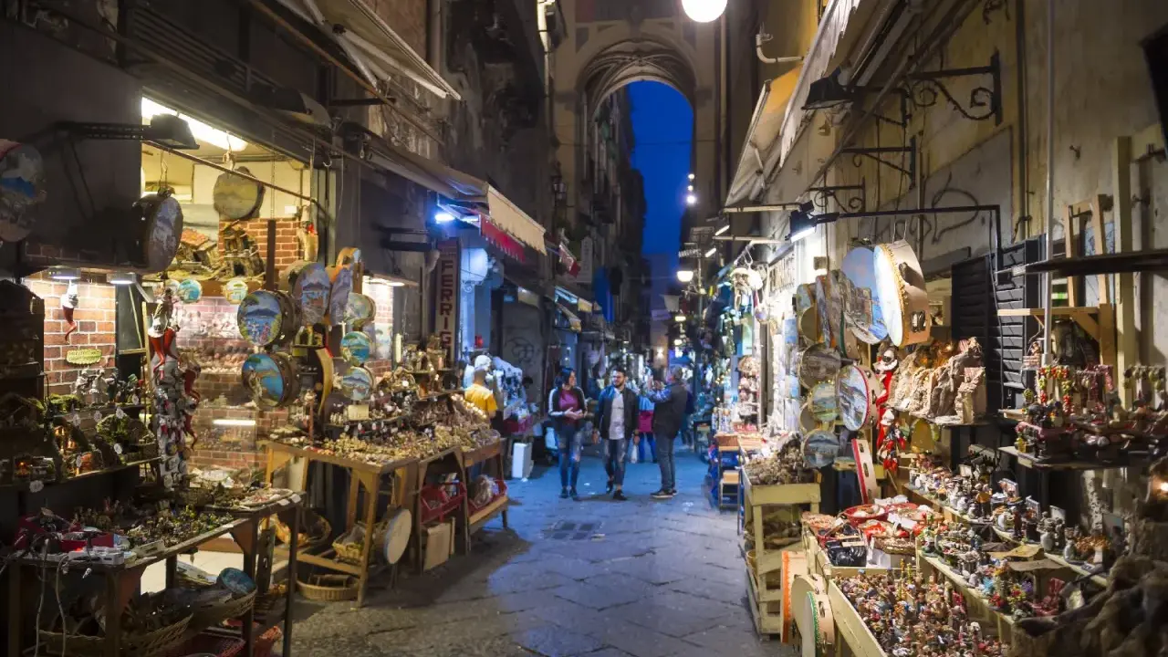 La via di San Gregorio Armeno, Napoli, illuminata di sera, con botteghe artigiane piene di presepi e statuine.
