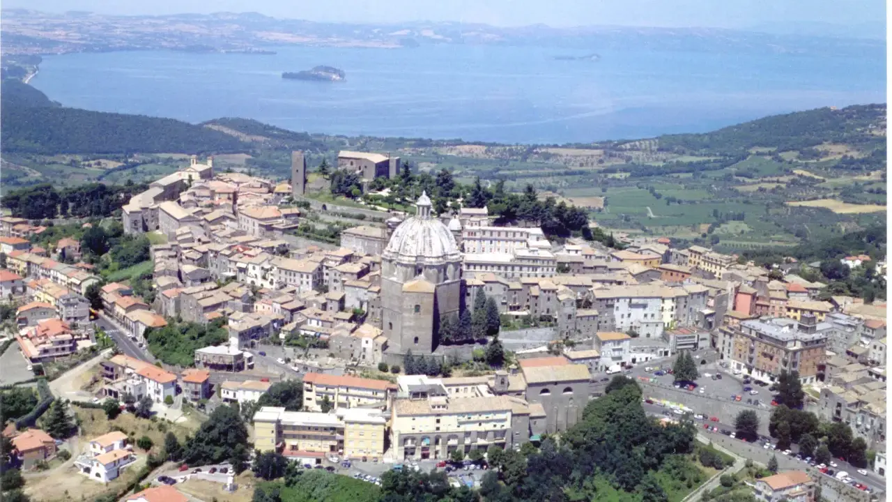 Vista aerea di un borgo italiano con cupola imponente, circondato da case con tetti rossi e affacciato su un lago. Est est est.