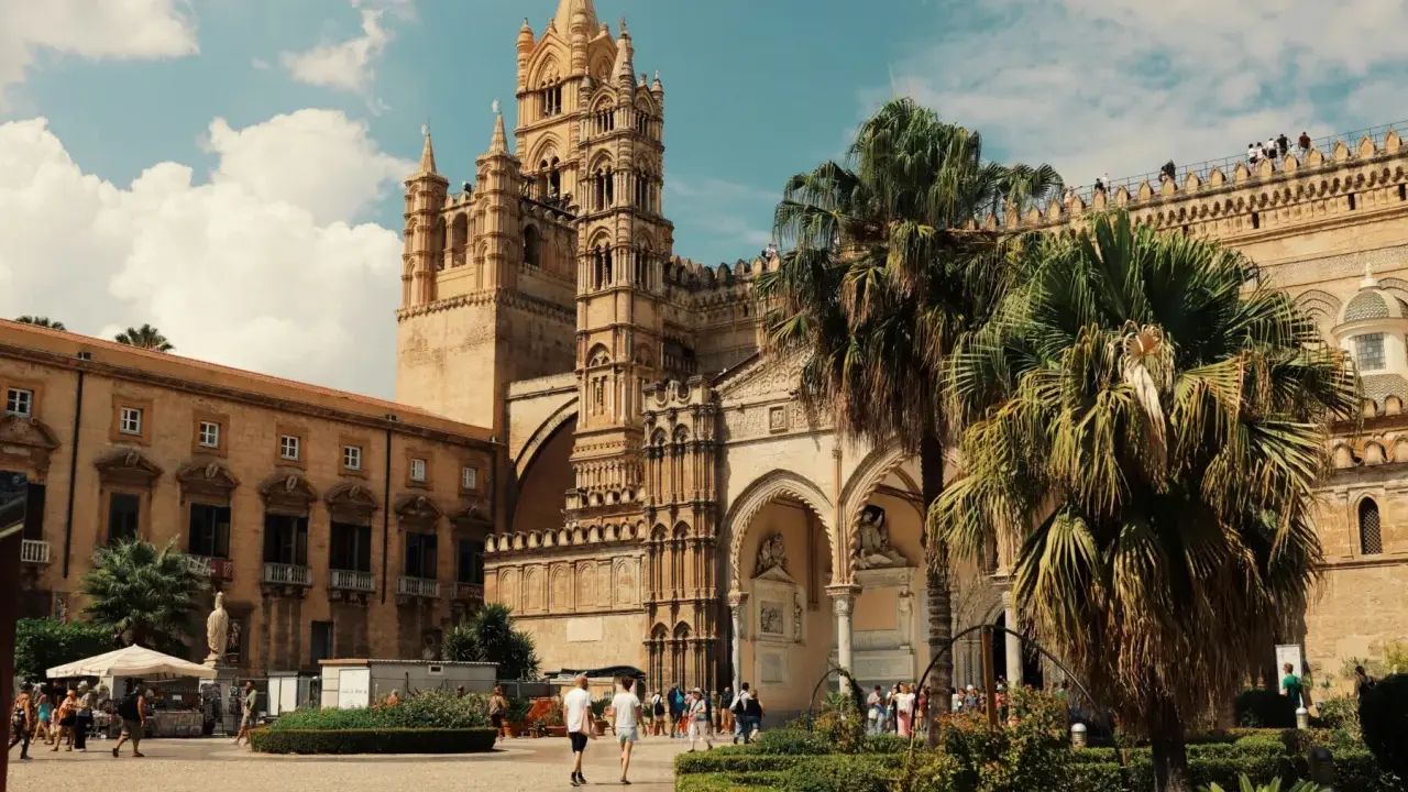 La Cattedrale di Palermo, un capolavoro di stili architettonici diversi, si erge maestosa sotto un cielo azzurro con palme rigogliose.