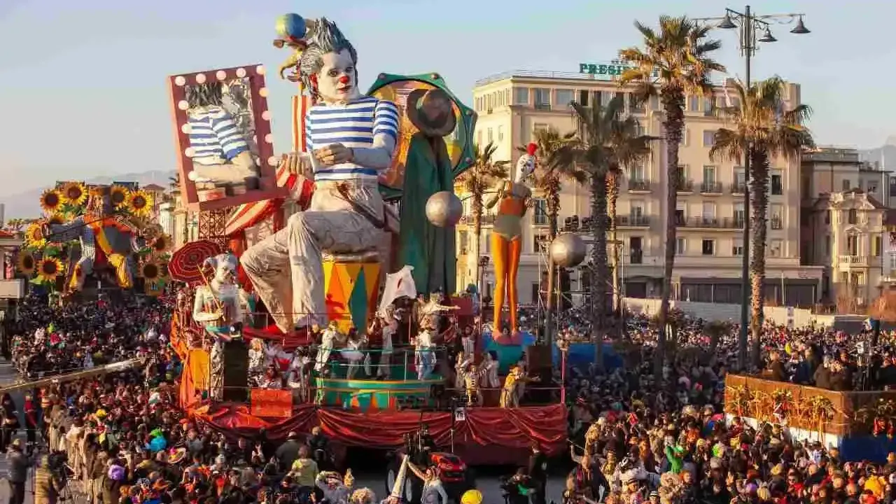 Carri allegorici e folla festante celebrano quando è Carnevale in Italia. Un clown gigante domina la scena, circondato da figure colorate e palme.
