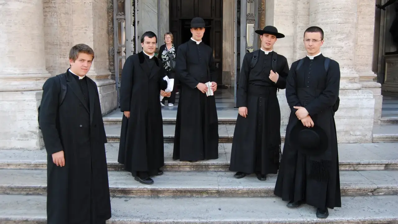 Cinque giovani seminaristi in abiti talari neri, alcuni con cappello, posano sulle scale di un edificio storico.