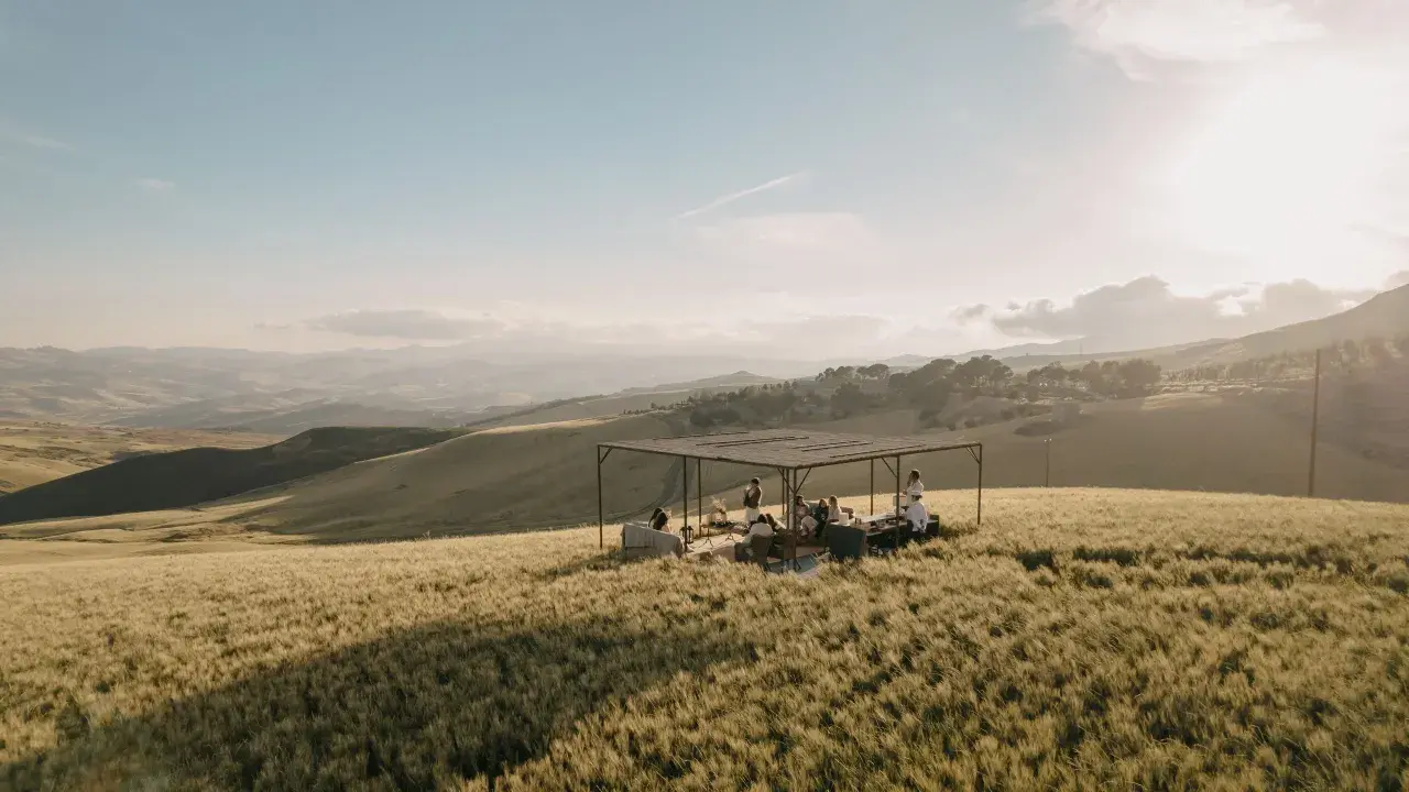 Un gruppo di persone si riunisce sotto una tettoia in un campo di grano dorato, godendosi la tipica bellezza siciliana al tramonto.