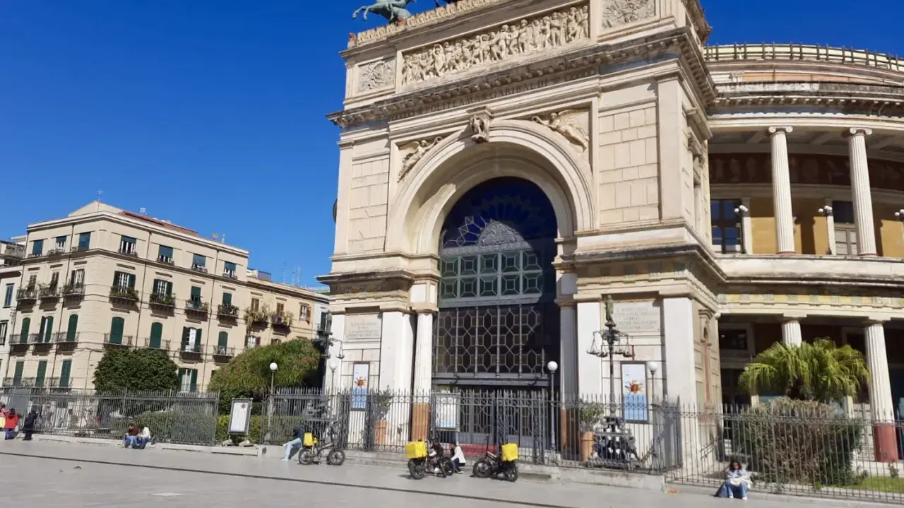 Il **Teatro Politeama Garibaldi** di Palermo, con la sua imponente facciata neoclassica, si staglia sotto un cielo azzurro. Tre scooter elettrici gialli sono parcheggiati davanti.