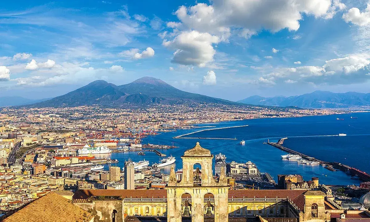 Vista panoramica di Napoli città con il Vesuvio sullo sfondo, il porto e un campanile in primo piano.