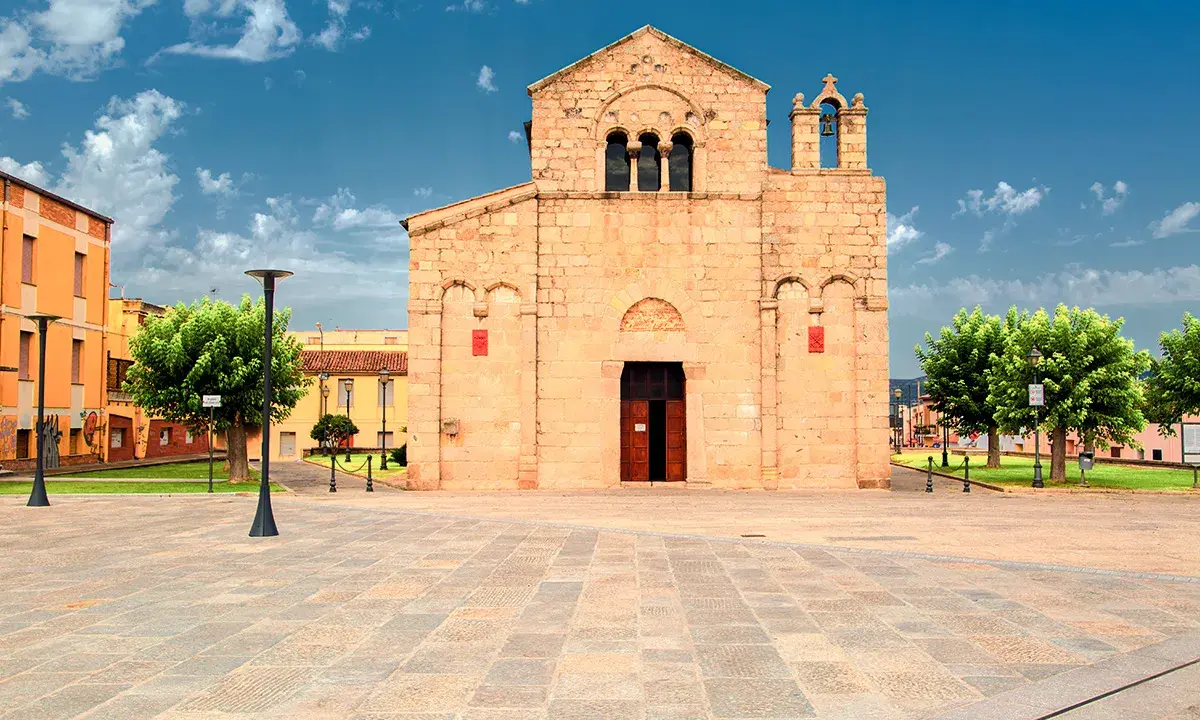 Piazza del centro di Olbia con la Basilica di San Simplicio, un'antica chiesa romanica in pietra chiara, sotto un cielo azzurro con nuvole bianche.