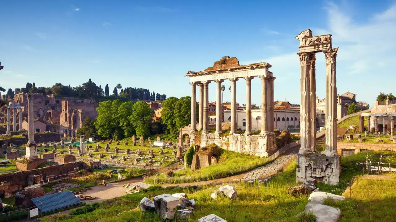 Antiche rovine del Foro Romano illuminate dal sole, con colonne imponenti e resti di templi sotto un cielo azzurro.