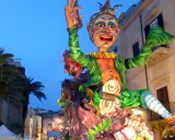 Carro allegorico con maschere di carnevale siciliane, un personaggio gioioso con vestiti verdi e arancioni sfila tra palme e palazzi illuminati.