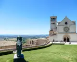 Statua equestre domina il prato verde di Assisi. Cosa vedere: la Basilica di San Francesco, con la sua imponente facciata e campanile, si staglia contro il cielo azzurro.