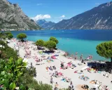 Spiaggia di Limone sul Garda affollata di bagnanti sotto le montagne. Acqua cristallina e sole splendente.