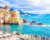 Case colorate a picco sul mare turchese, barche ormeggiate e un promontorio roccioso: un angolo di Genova che ricorda le sue spiagge più belle.