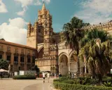 La Cattedrale di Palermo, un capolavoro di stili architettonici diversi, si erge maestosa sotto un cielo azzurro con palme rigogliose.