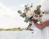 Un bouquet di rose e peonie, come quello che vorrei per a mia moglie, tenuto da una sposa con un abito bianco, con un lago sullo sfondo.