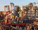 Carri allegorici e folla festante celebrano quando è Carnevale in Italia. Un clown gigante domina la scena, circondato da figure colorate e palme.