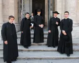 Cinque giovani seminaristi in abiti talari neri, alcuni con cappello, posano sulle scale di un edificio storico.