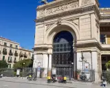 Il **Teatro Politeama Garibaldi** di Palermo, con la sua imponente facciata neoclassica, si staglia sotto un cielo azzurro. Tre scooter elettrici gialli sono parcheggiati davanti.