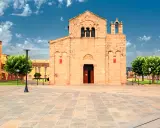 Piazza del centro di Olbia con la Basilica di San Simplicio, un'antica chiesa romanica in pietra chiara, sotto un cielo azzurro con nuvole bianche.