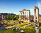 Antiche rovine del Foro Romano illuminate dal sole, con colonne imponenti e resti di templi sotto un cielo azzurro.
