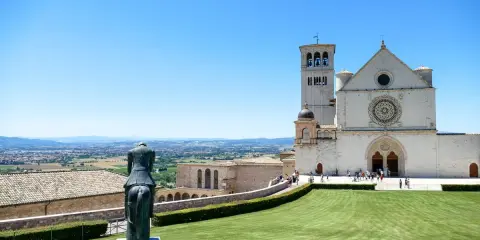 Statua equestre domina il prato verde di Assisi. Cosa vedere: la Basilica di San Francesco, con la sua imponente facciata e campanile, si staglia contro il cielo azzurro.