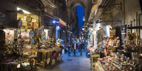 La via di San Gregorio Armeno, Napoli, illuminata di sera, con botteghe artigiane piene di presepi e statuine.