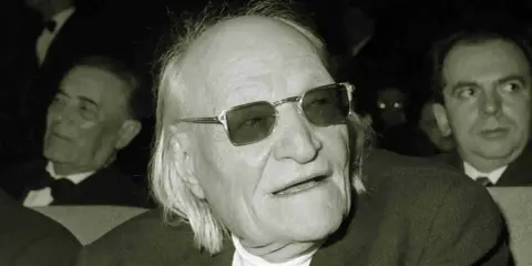 Un uomo anziano con occhiali da sole, capelli lunghi e bianchi, sembra un poeta. Sono una creatura ungaretti.