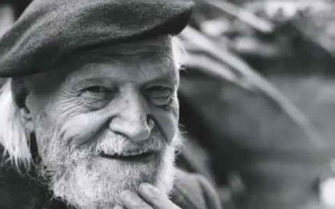 Un anziano con barba e berretto, il suo sorriso saggio evoca il significato che la morte si sconta vivendo appieno ogni istante.