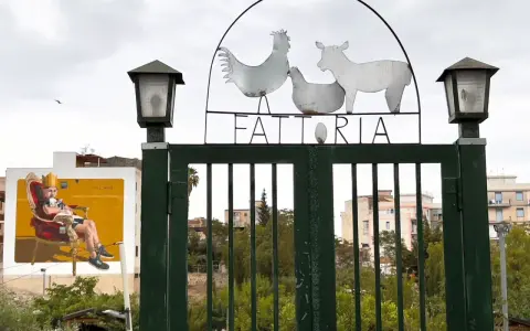Cancello verde con insegna "FATTORIA" e sagome di animali. A sinistra, murale di un uomo su trono. Danisinni, Palermo.