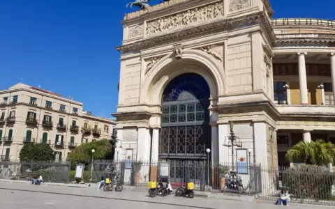 Il **Teatro Politeama Garibaldi** di Palermo, con la sua imponente facciata neoclassica, si staglia sotto un cielo azzurro. Tre scooter elettrici gialli sono parcheggiati davanti.