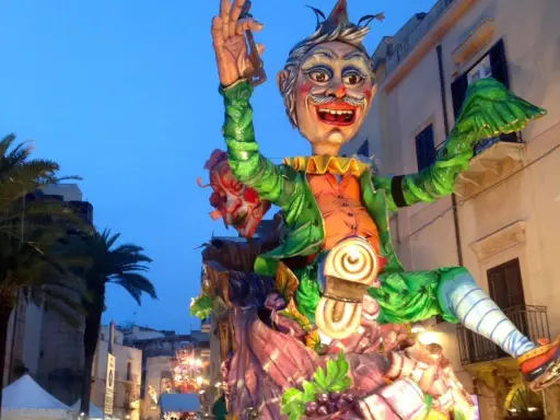 Carro allegorico con maschere di carnevale siciliane, un personaggio gioioso con vestiti verdi e arancioni sfila tra palme e palazzi illuminati.