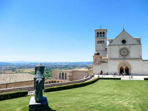 Statua equestre domina il prato verde di Assisi. Cosa vedere: la Basilica di San Francesco, con la sua imponente facciata e campanile, si staglia contro il cielo azzurro.