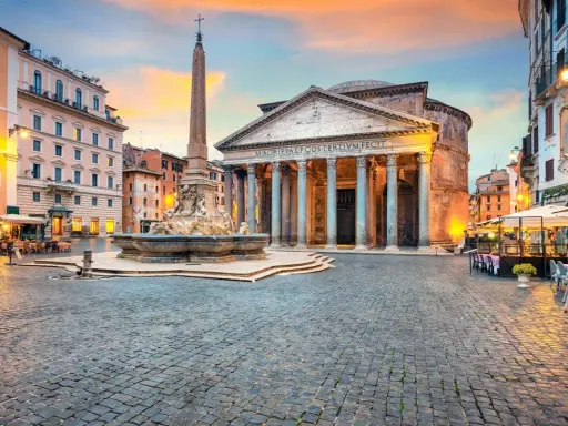 Piazza illuminata con il Pantheon, una chiesa maestosa, e una fontana con obelisco. Tavolini all'aperto invitano a godersi la sera romana.