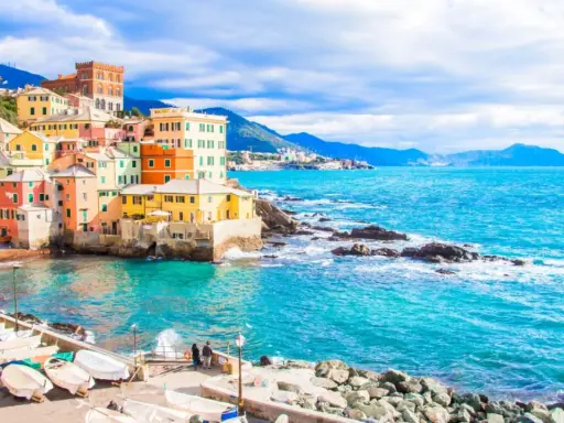 Case colorate a picco sul mare turchese, barche ormeggiate e un promontorio roccioso: un angolo di Genova che ricorda le sue spiagge più belle.