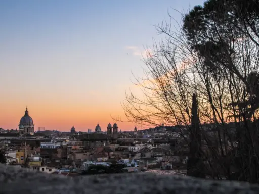 Un magico pincio roma tramonto con cupole che si stagliano contro il cielo aranciato, alberi spogli in primo piano.