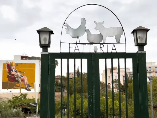 Cancello verde con insegna "FATTORIA" e sagome di animali. A sinistra, murale di un uomo su trono. Danisinni, Palermo.