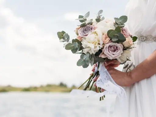 Un bouquet di rose e peonie, come quello che vorrei per a mia moglie, tenuto da una sposa con un abito bianco, con un lago sullo sfondo.