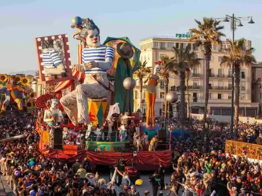 Carri allegorici e folla festante celebrano quando è Carnevale in Italia. Un clown gigante domina la scena, circondato da figure colorate e palme.