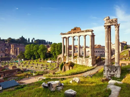 Antiche rovine del Foro Romano illuminate dal sole, con colonne imponenti e resti di templi sotto un cielo azzurro.