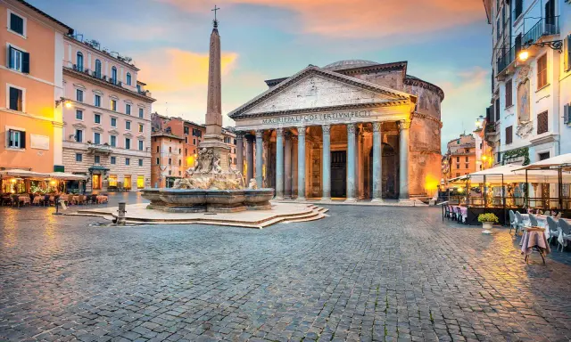 Piazza illuminata con il Pantheon, una chiesa maestosa, e una fontana con obelisco. Tavolini all'aperto invitano a godersi la sera romana.
