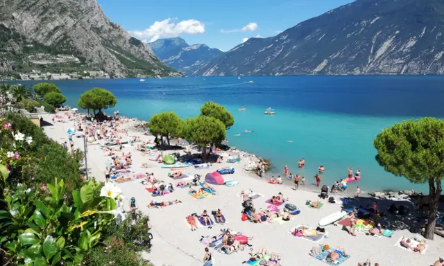 Spiaggia di Limone sul Garda affollata di bagnanti sotto le montagne. Acqua cristallina e sole splendente.