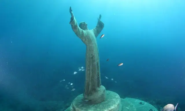 Il Cristo degli Abissi, una statua sommersa, veglia sul fondale blu, circondato da banchi di pesci che nuotano sereni.