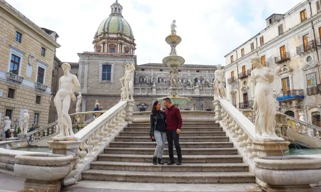 Coppia felice davanti alla Fontana Pretoria, un luogo imperdibile da vedere a Palermo, con le sue statue e la cupola in lontananza.