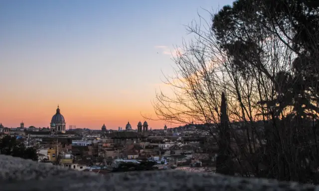 Un magico pincio roma tramonto con cupole che si stagliano contro il cielo aranciato, alberi spogli in primo piano.
