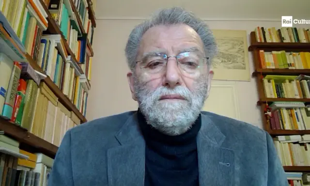 Uomo anziano con barba grigia, occhiali e giacca, circondato da scaffali pieni di libri. Il suo sguardo intenso suggerisce che ei fu significato.