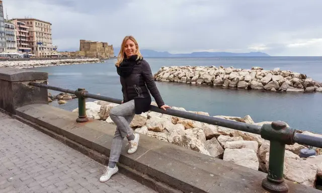 Donna sorridente appoggiata a un parapetto con Castel dell'Ovo sullo sfondo, una giornata tipica di napoli a gennaio.