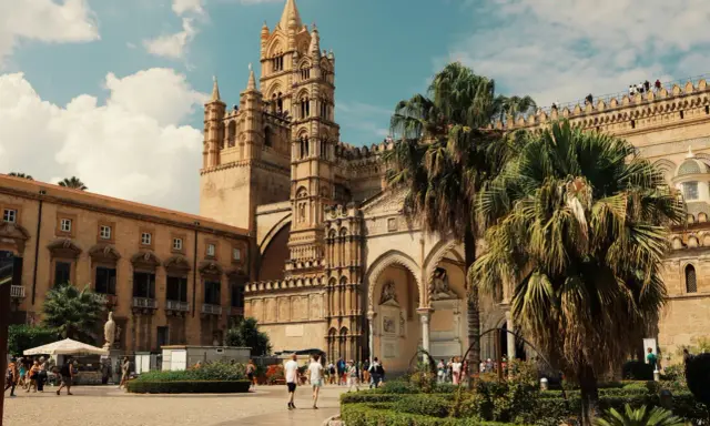 La Cattedrale di Palermo, un capolavoro di stili architettonici diversi, si erge maestosa sotto un cielo azzurro con palme rigogliose.