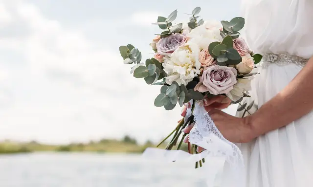 Un bouquet di rose e peonie, come quello che vorrei per a mia moglie, tenuto da una sposa con un abito bianco, con un lago sullo sfondo.